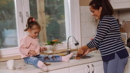 Mama pere sudove dok ćerka sedi pored