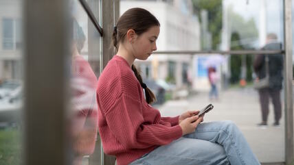 Devojčica na autobuskom stajalištu sedi i gleda u telefon